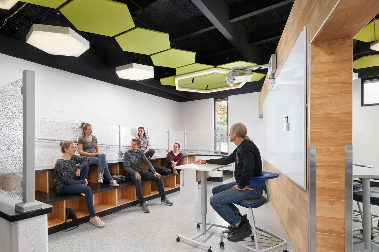 Male teacher sitting at movable desk teaching several students in modern classroom with green tiles on the ceiling