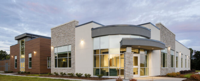 Modern building with a mix of brick and glass exteriors, featuring a curved entrance under a cloudy sky. Lush green lawn in the foreground at Jonas Hill Hospital and Behavioral Health Clinic.