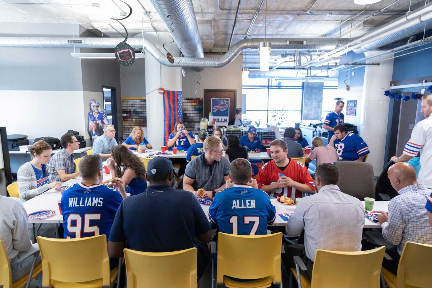 CPL's Buffalo NY team sits around tables wearing Buffalo Bills jerseys and talking to each other. Image shows mostly men sitting around tables. 