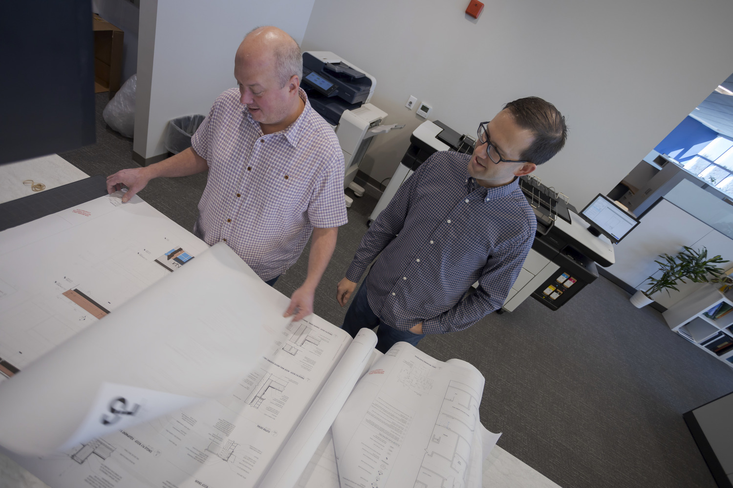 Image shows two white men, one older and wearing a plaid button-up shirt, the other is younger and wearing glasses and a blue button-up shirt reading blueprints at CPL Pittsburgh.