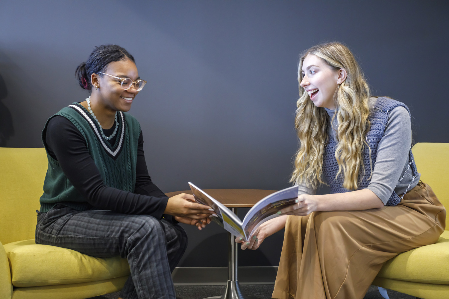 Image shows two women at CPL Pittsburgh, one is a young black woman with her hair pulled back wearing glasses and a green top sitting in a yellow chair next to a young white woman with long blonde hair wearing a gray top and mustard color pants. The two are having a conversation about the book the blonde woman is holding.