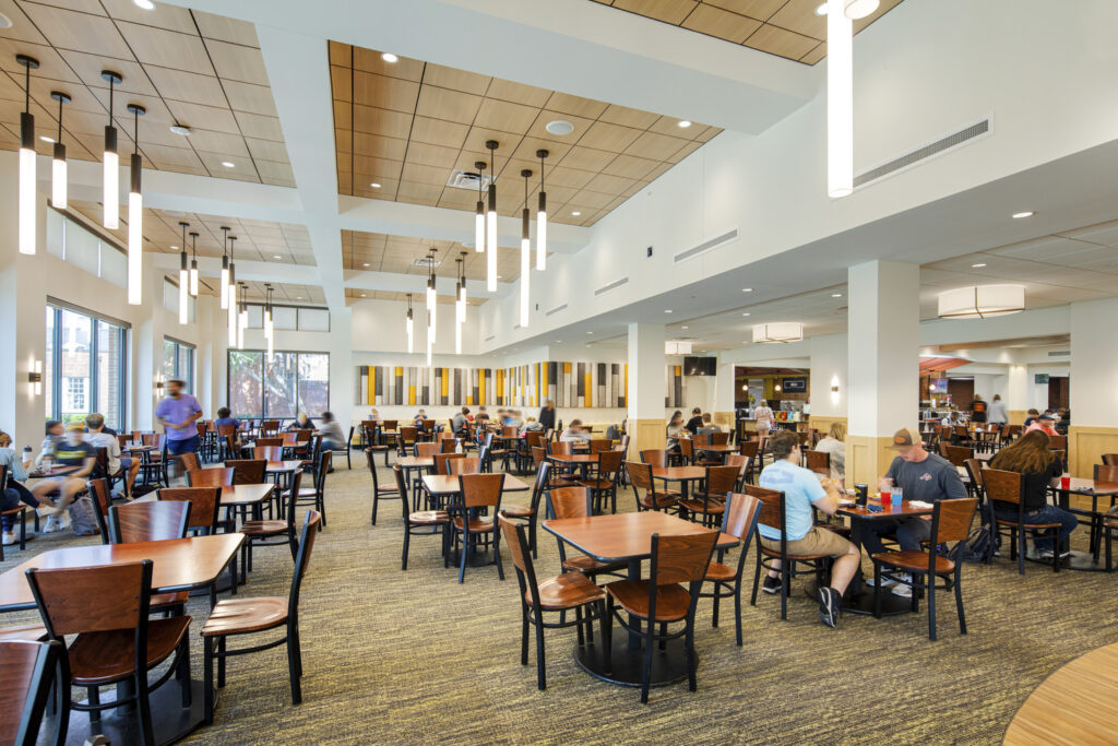 Image shows a modern dining hall with hanging lighting and many tables surrounded by chairs. Students are sitting at the tables together and eating food. This is the Randolph-Macon College Estes Dining Hall.