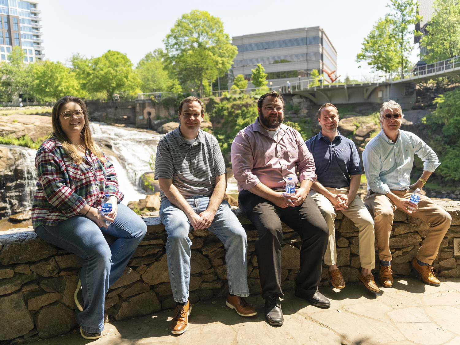 Image shows a group of 5 white people sitting on a small fence made of rocks in front of a water fall. They are all smiling. These are the members of CPL Greenville.