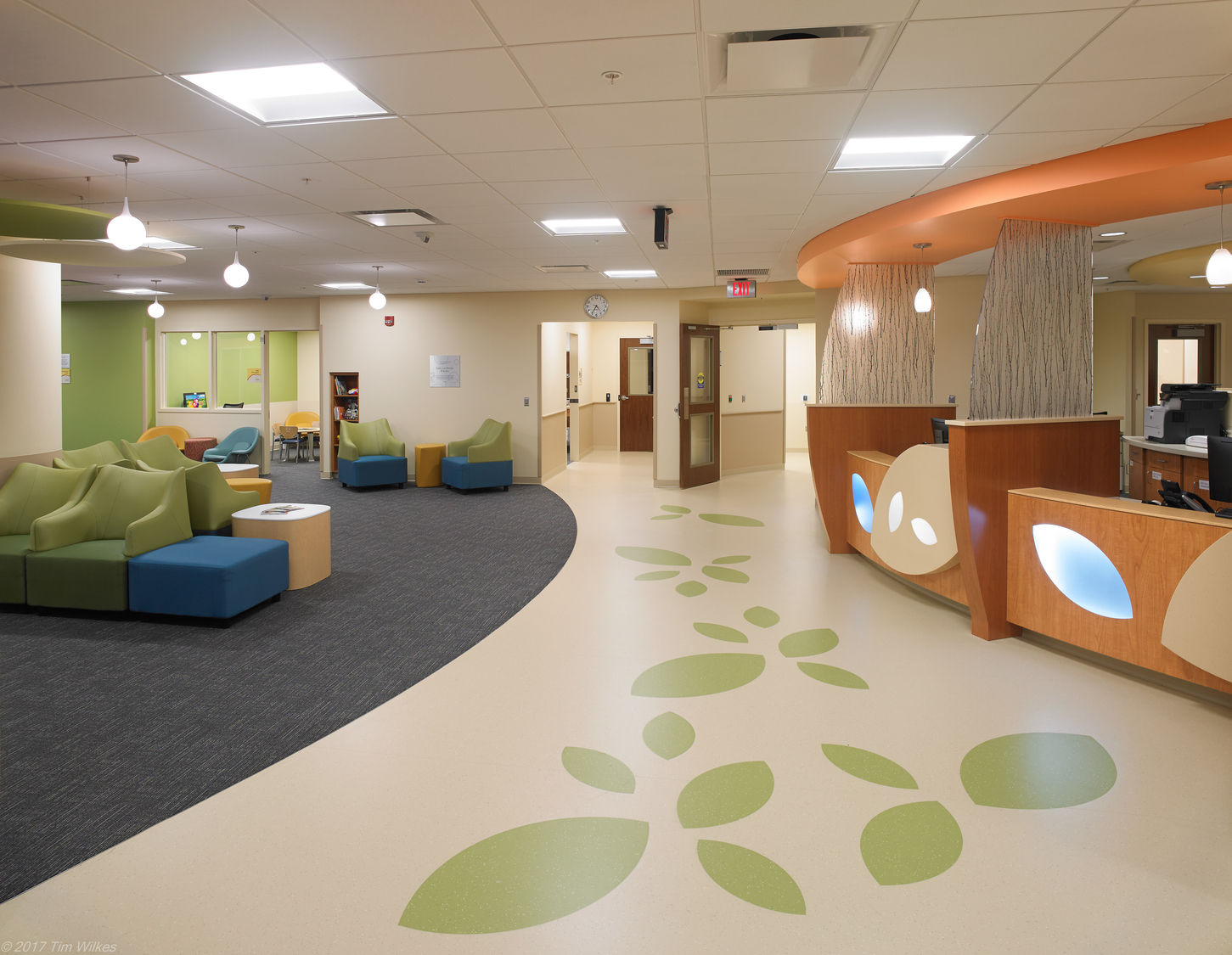 Image shows a fun lobby design that has leaves on the walkway, green comfortable seating, and desks for people to speak to the receptionist. This space is The University of Rochester Medical Center’s (URMC) Levine Autism Clinic.