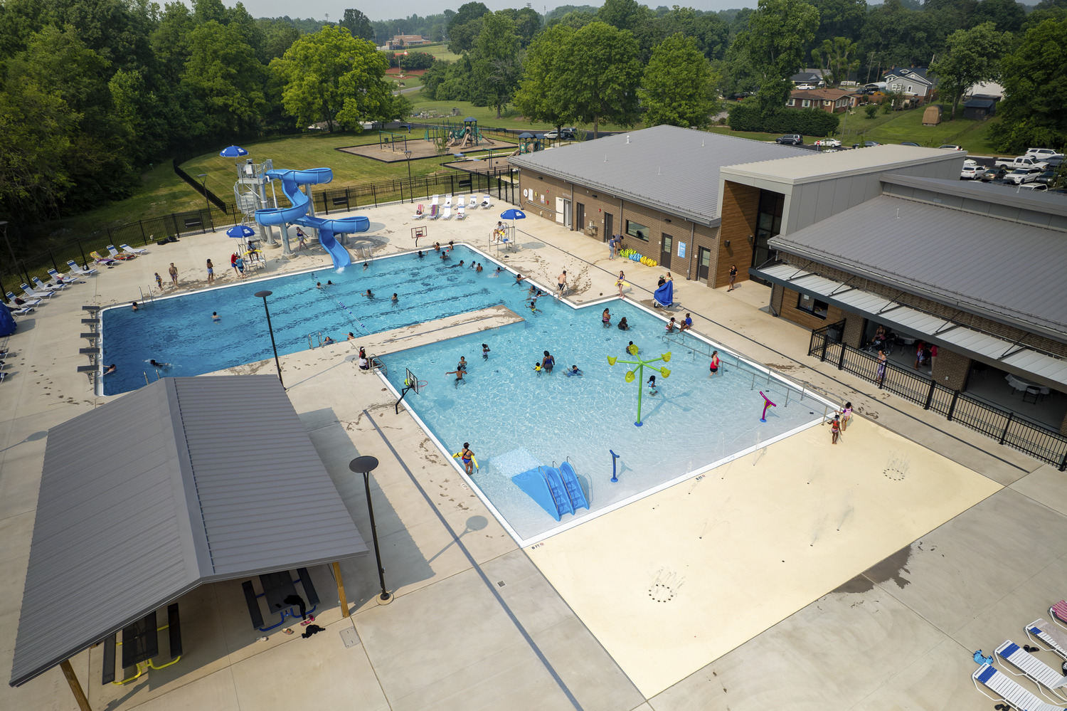 Image shows the pools and community center at the Thomasville Aquatic and Community Center. A playground can be seen in the background. There are people playing in the pool.