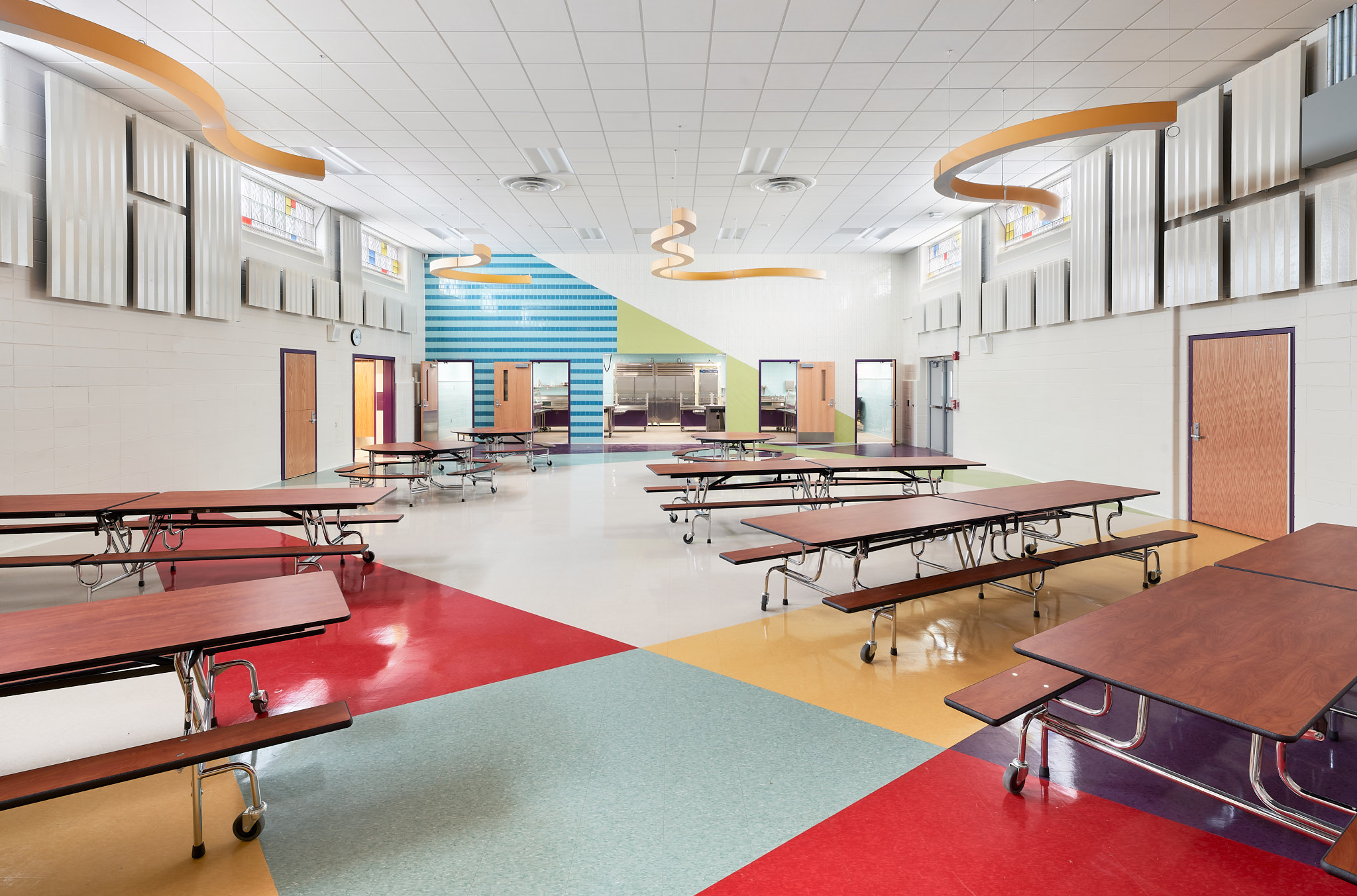 A modern and colorful cafeteria is shown. There are multiple tables. The flooring has red, blue, yellow, and white/gray. There are interesting hanging features as well.