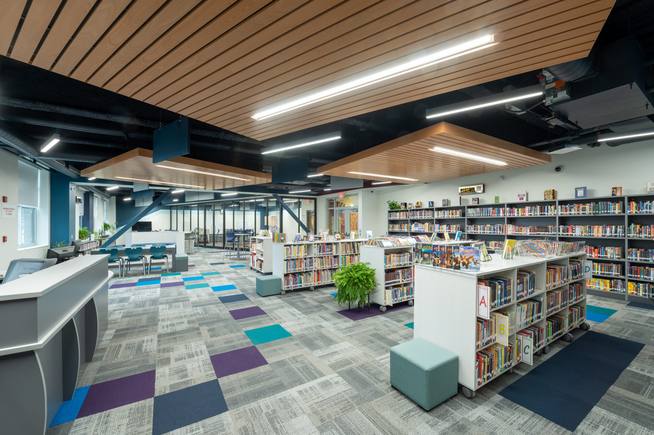 The media center at Avoca CSD is shown. The floor has colorful carpet that is gray with turquiose, purple, and navy elements. There are book shelves and places to sit. 