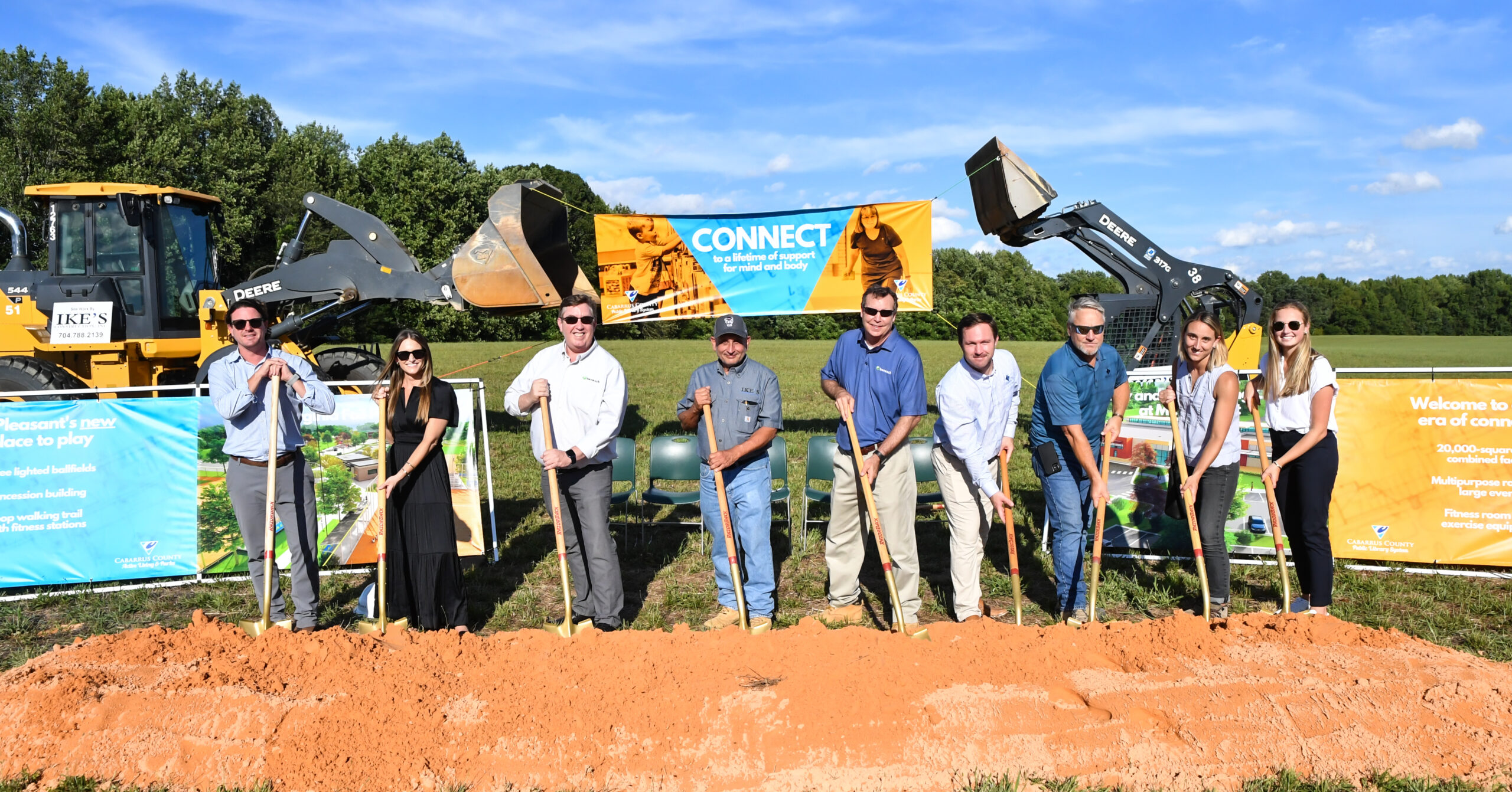 Image shows a group of people holding shovels and digging into the dirt. Behind them are two construction vehicles with their buckets up. The sky is blue in the background.