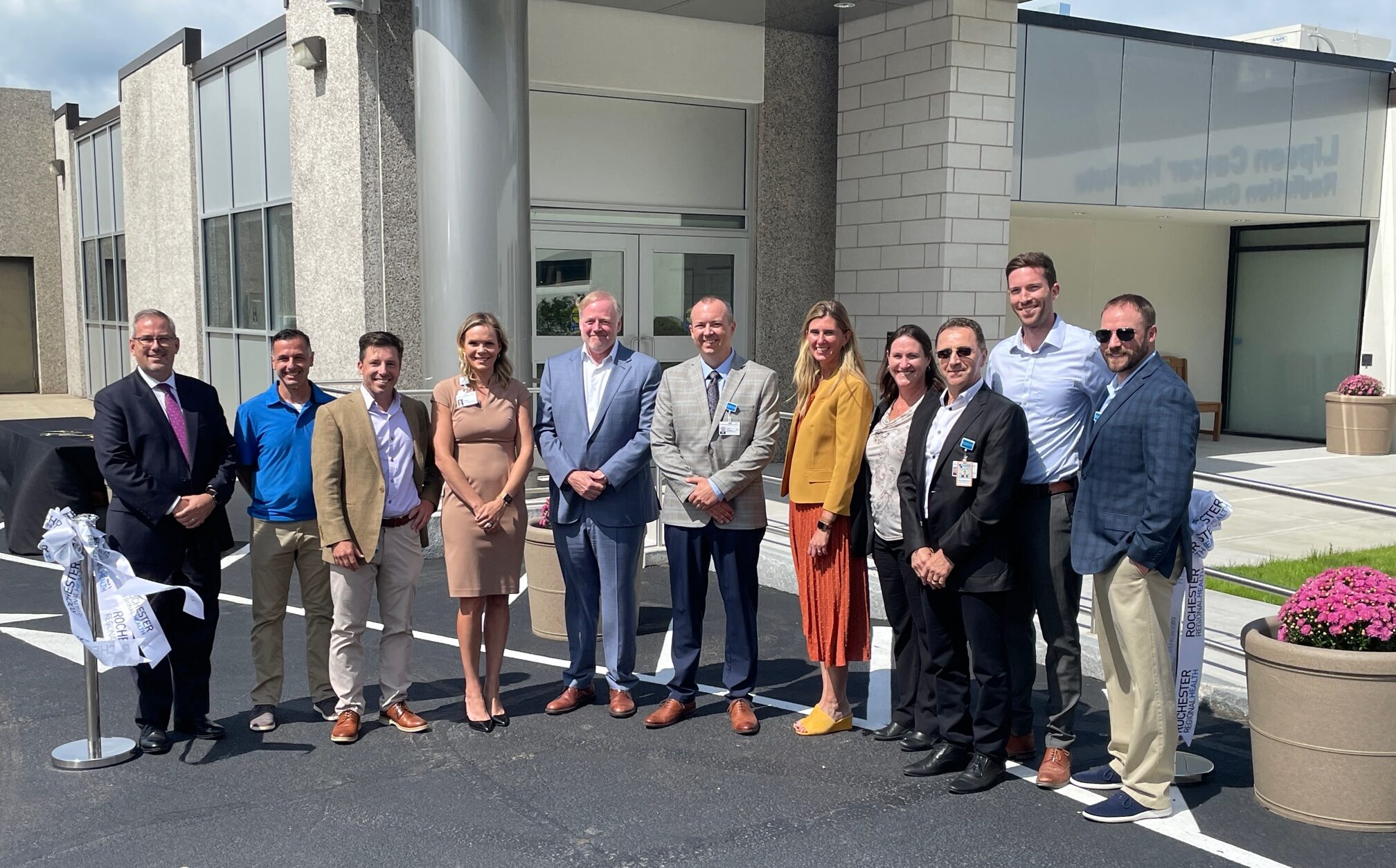 A group of 11 people, dressed in business clothes stands in front of a brick building, The Clifton Springs Hospital Enhanced Emergency Department. Posts with ribbons stand next to the group.