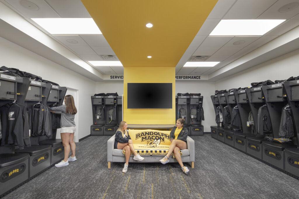 A locker room is shown with gray cubbies with personal items in them. Down the middle of the room is a gold stripe and a couch that reads "Randolph-Macon" Two women sit on the couch while one woman is at one of the cubbies.