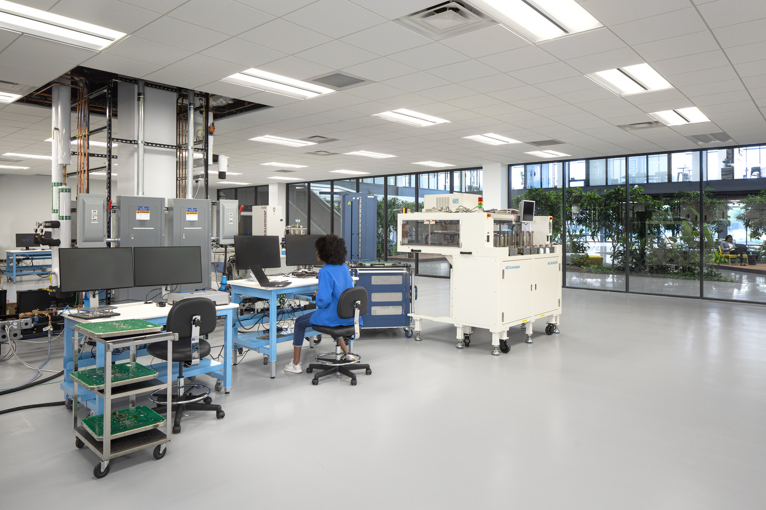 A large room with windows that show indoor plants. In the center there's lab equipment and a woman sits at a desk. This lab is at Analog Devices.