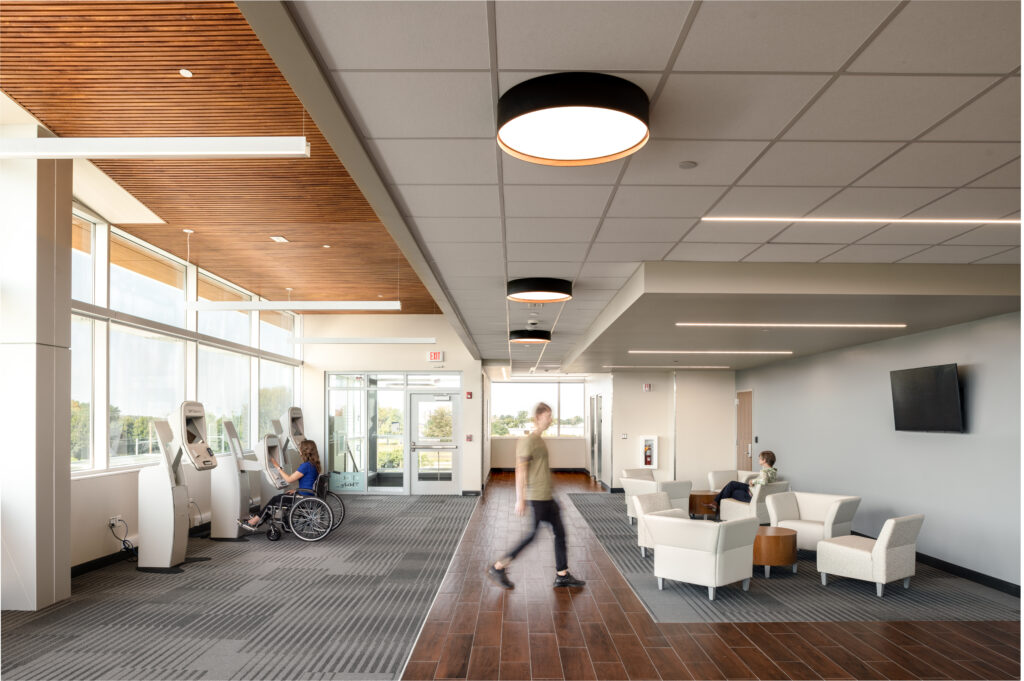 R21_16060_00 A waiting room at RRH Batavia Medical Destination Center with wood floors and grey carpeting. There are round light fixtures on the ceiling. A woman in a blue shirt in a wheelchair uses a self-checkin kiosk, a man walks, and a woman sits in a white armchair.