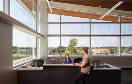A receptionist in a blue shirt talks to a man in a tan shirt at RRH Batavia Medical Destination Center. There are windows on all of the walls, and a wood paneled ceiling.