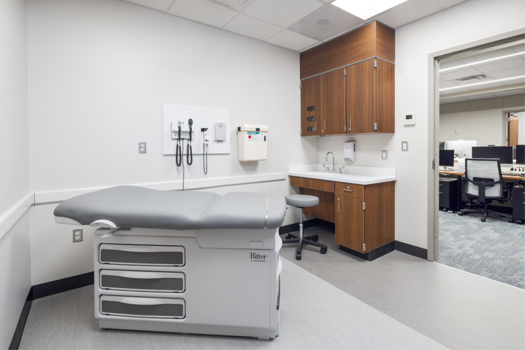 R21_16060_00_Rochester_Regional_Health_N8 Medical exam room at RRH Batavia Medical Destination Center with grey floors and white walls, grey bed, and wood cabinets. Medical equipment on the wall, and a view through the doorway to a computer station.