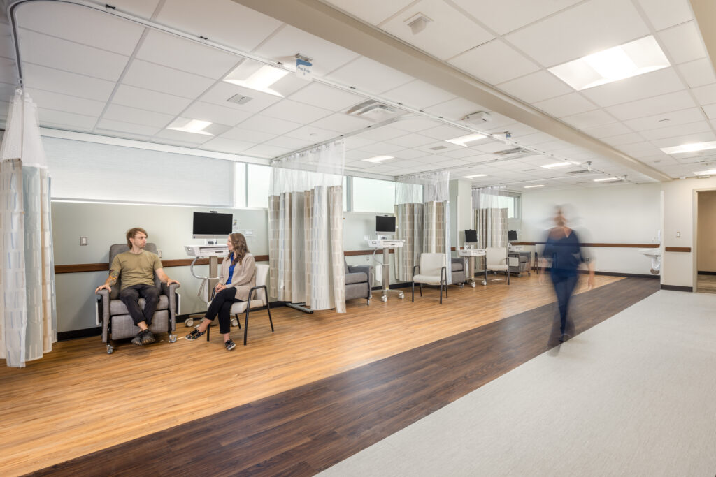 R21_16060_00_Rochester_Regional_Health_N9 A medical treatment area at RRH Batavia Medical Destination Center. A man in a tan shirt with tattoos sits in a treatment chair as a female doctor sits next to him. A nurse walks across the front area past the curtained off treatment stations.
