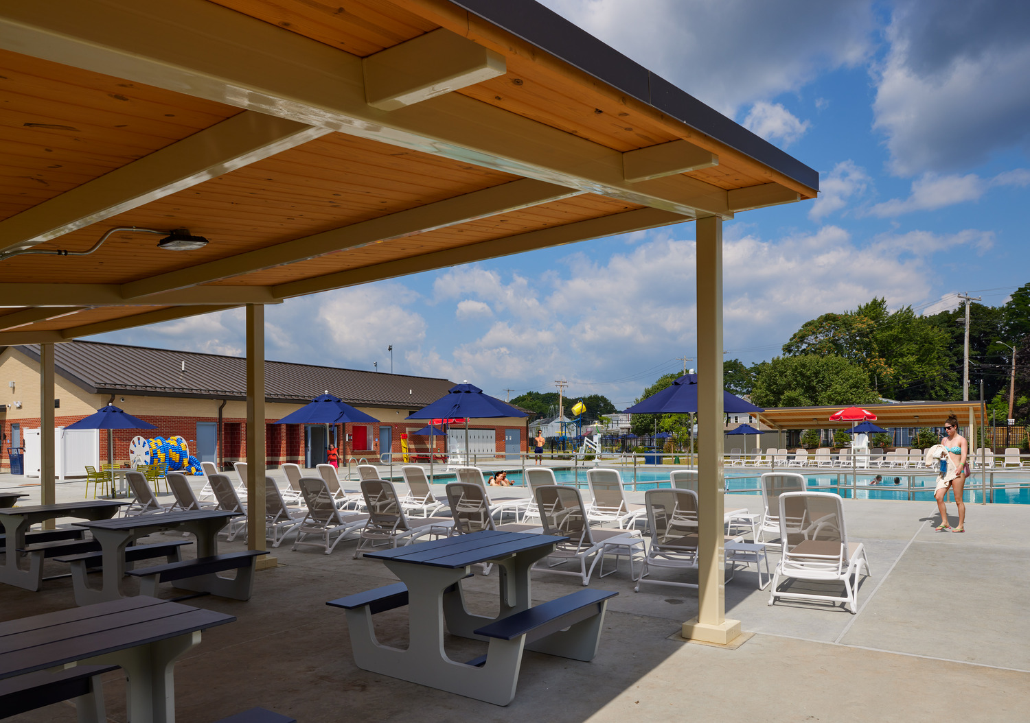 Covered patio area at a poolside with picnic tables and white lounge chairs facing a blue pool at Delano-Hitch Aquatic Center in Newburgh, NY. People swim under blue umbrellas, trees in the background.
