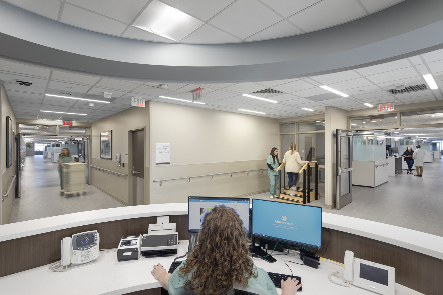 Atrium Health Pineville ICU desk and hallway. A nurse can be seen at the desk and another in the hall helping a patient on stairs.