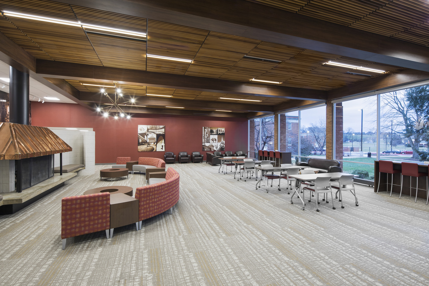 A modern university lounge space with a gray carpet; a large fireplace; ample seating areas; desks for studying; and red walls. 