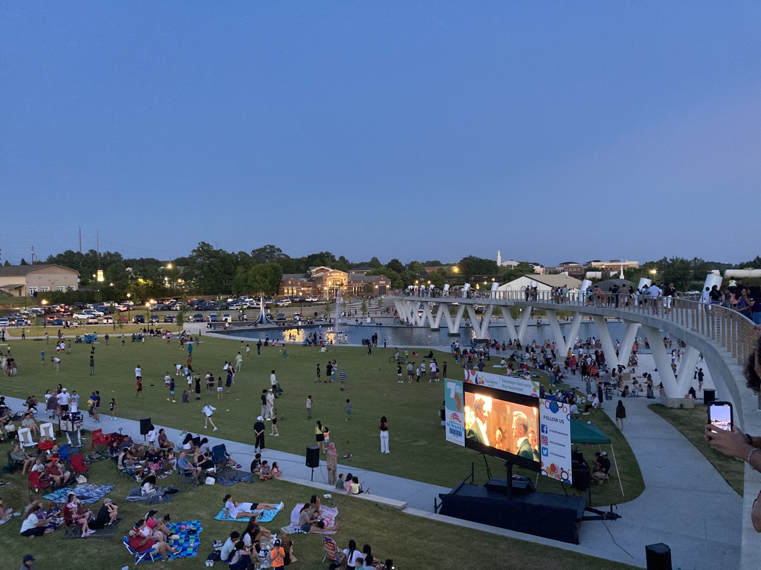 An aerial view of numerous people watching a movie in an expansive public park, with a long white bridge over water.