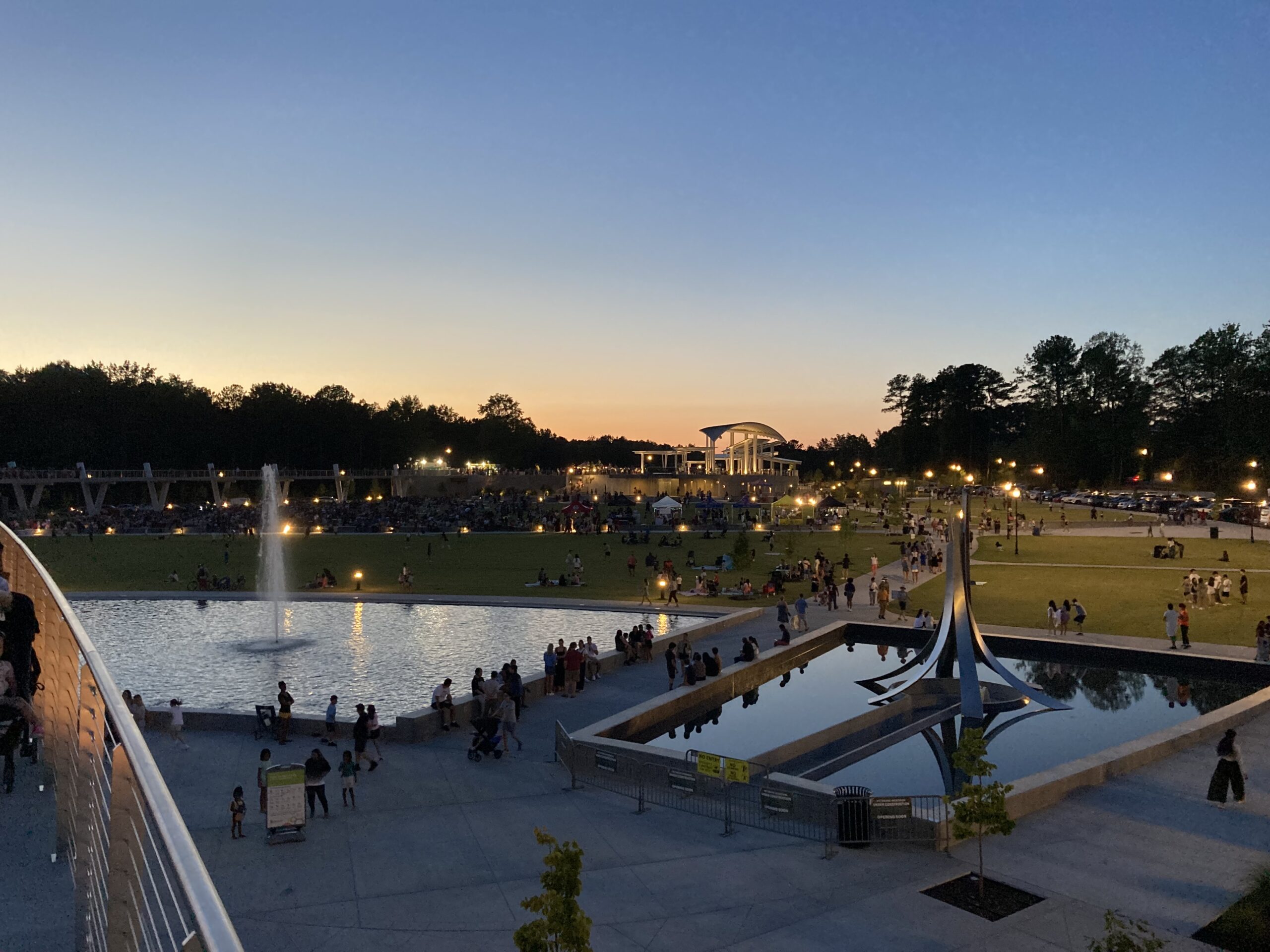 A picture of an expansive public park at dusk, with grassy lawns and water features, and a white canopy in the background.