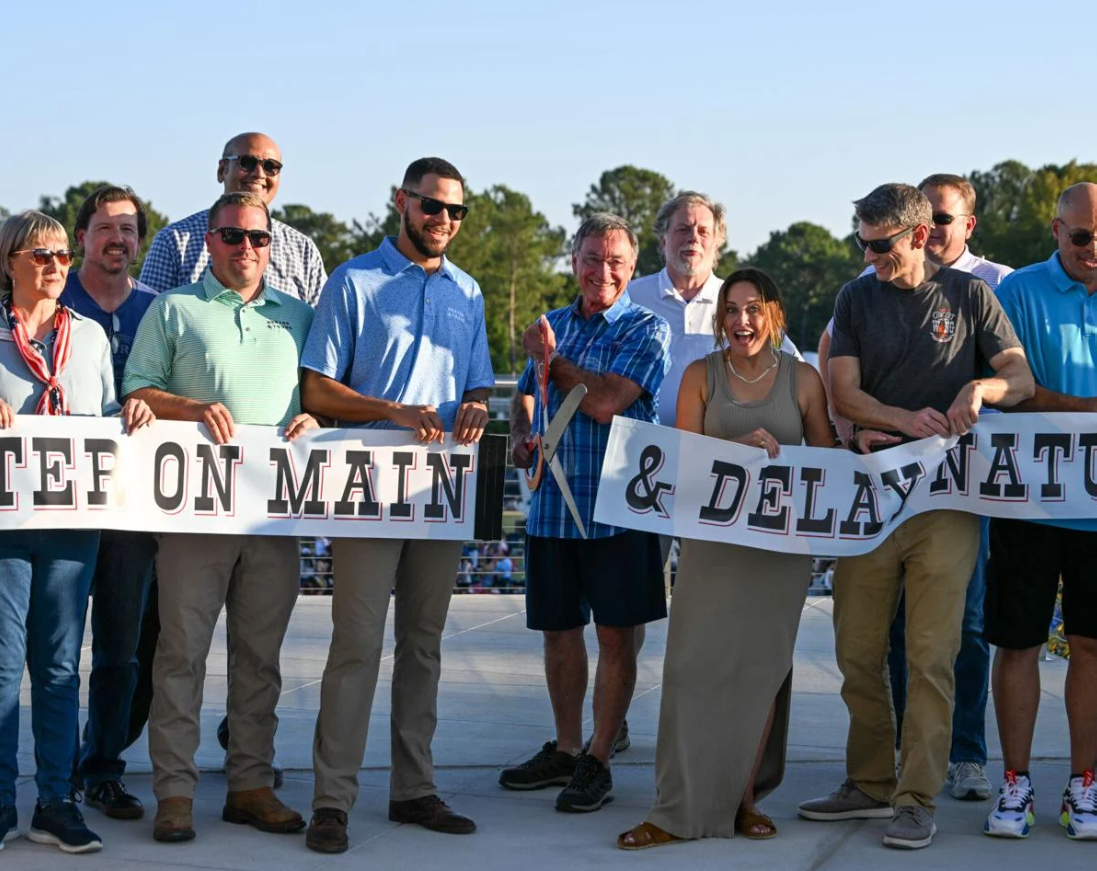 A group of people cutting the ribbon at a grand opening for a new park. A man in the middle holds a large pair of scissors and cuts the ribbon.