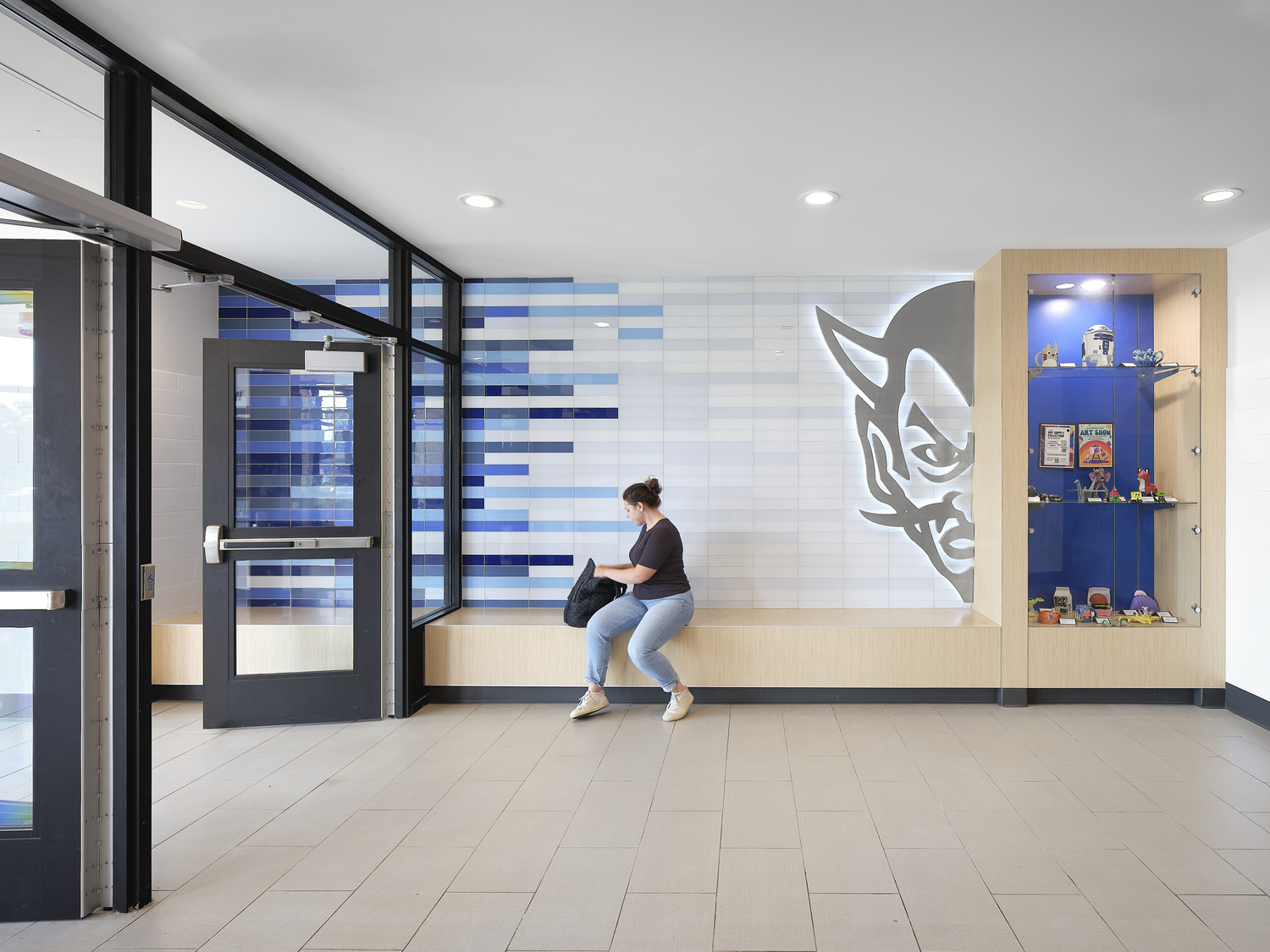 A student sits on a bench in a light and airy high school hallway, with blue tile details on the wall and a Blue Devil mascot prominently featured and backlit.