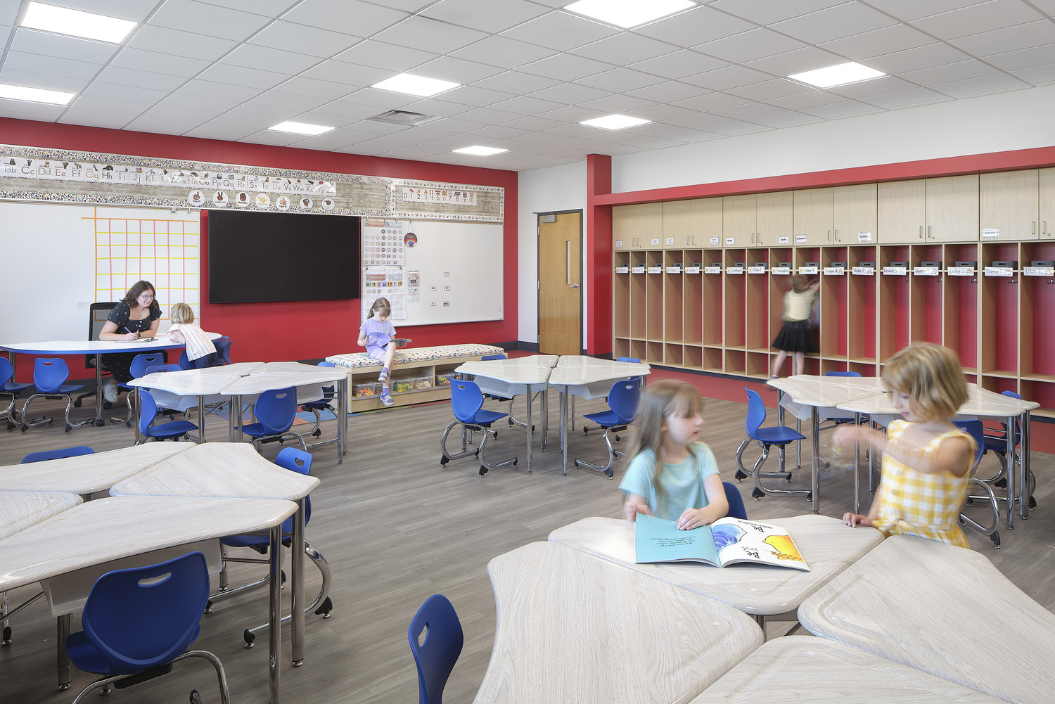 A red elementary school classroom, with a teacher sitting at the front and students sitting at their desks. There is a TV by the whiteboard.