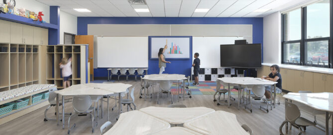 A Kenmore Tonawanda CSD modern classroom with light wood floors, round tables, and gray chairs. A teacher and student interact at the smartboard. Bookshelves line the walls.