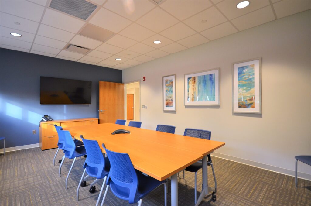 A conference room with a long wooden table, blue chairs, wall art, and a monitor at the head of the room.