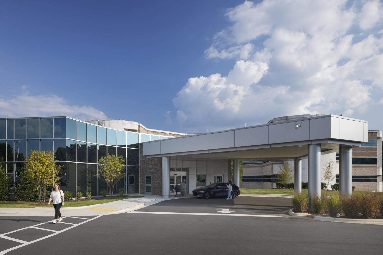 The exterior of a medical facility with a unique class facade, complemented by warm ledgestone. The entrance is a circular flow, allowing easy patient dropoffs. The background includes a blue sky with clouds.