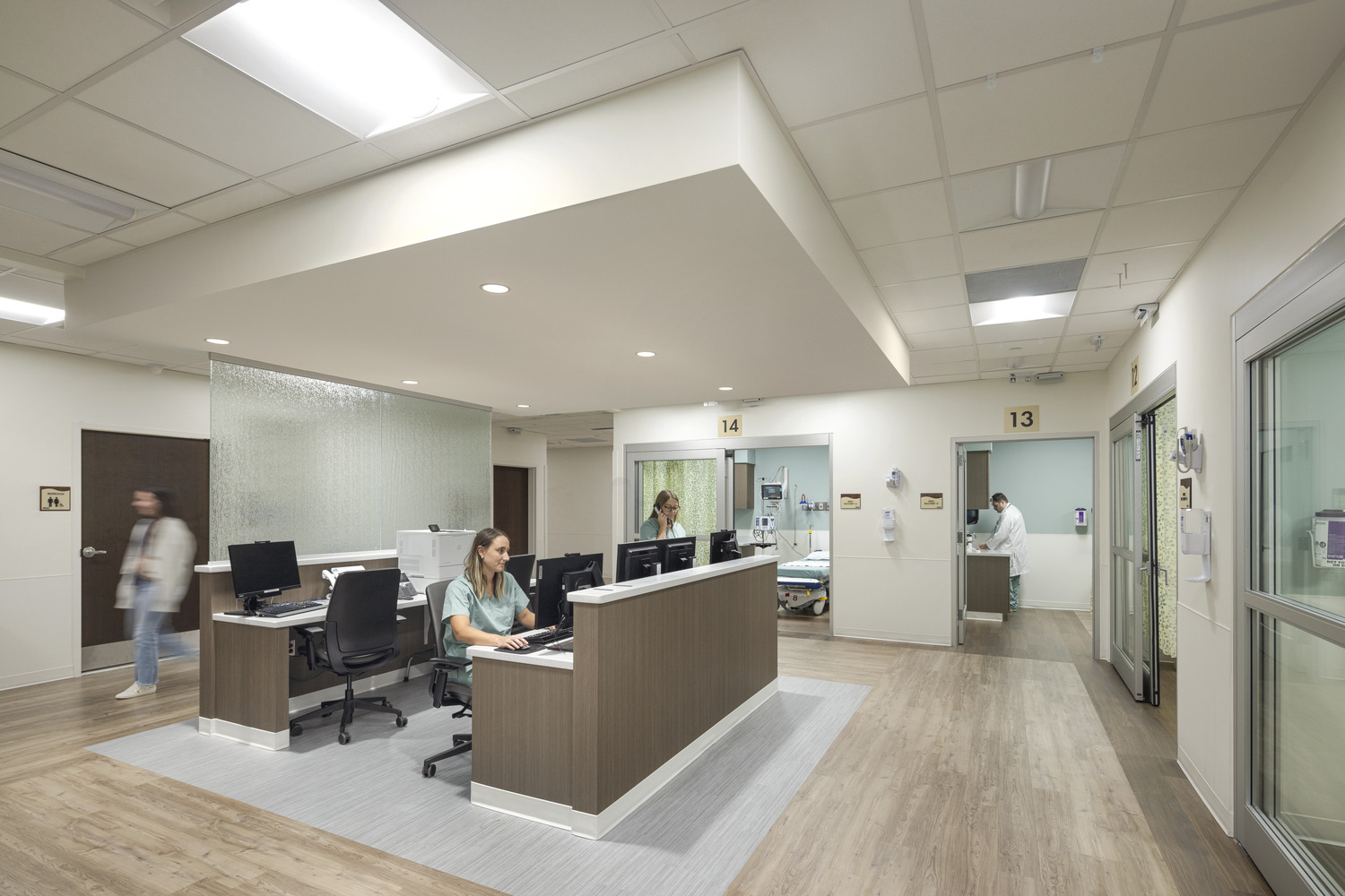 A centralized nursing station in a cardiac care facility with clear sightlines to patient rooms. A nurse in scrubs is pictured sitting at the front desk.