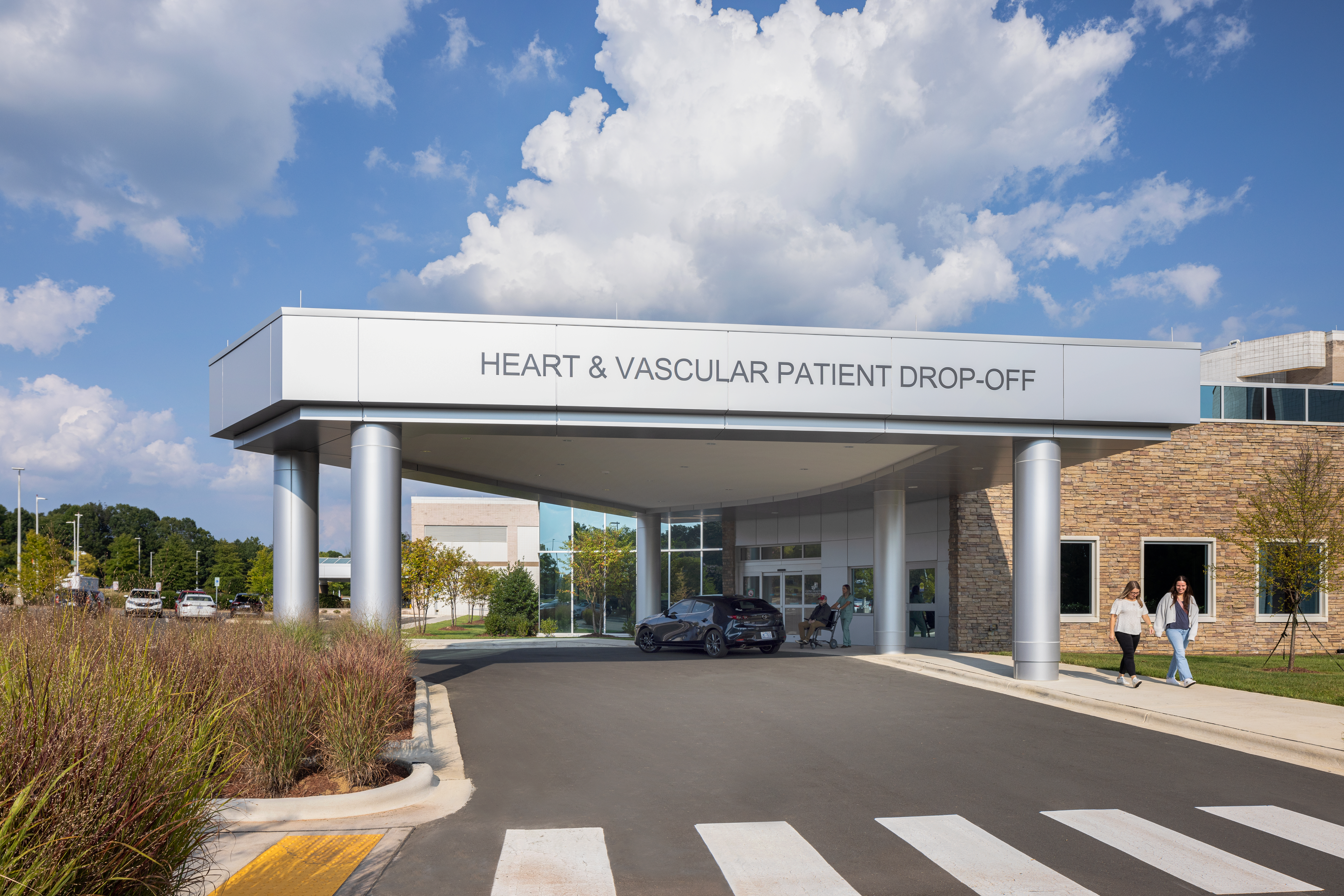 An exterior of a Heart & Vascular center for advanced cardiac care. Its entrance has a circular flow for easy drop-offs. The background includes a blue sky with clouds.