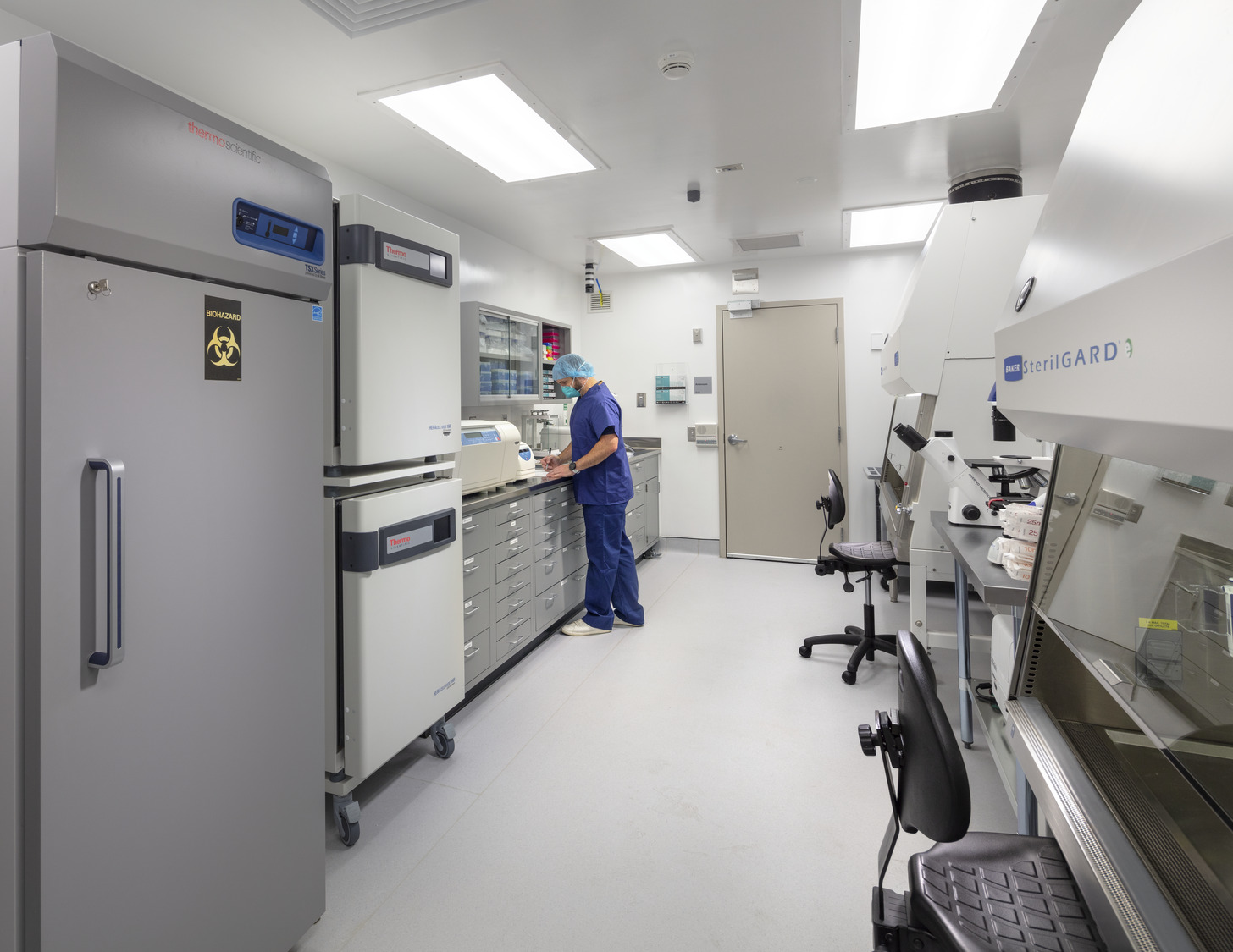 A researcher in a blue lab coat works in a higher education lab, equipped with advanced safety equipment including biosafety cabinets, refrigerators, and other lab instruments. 