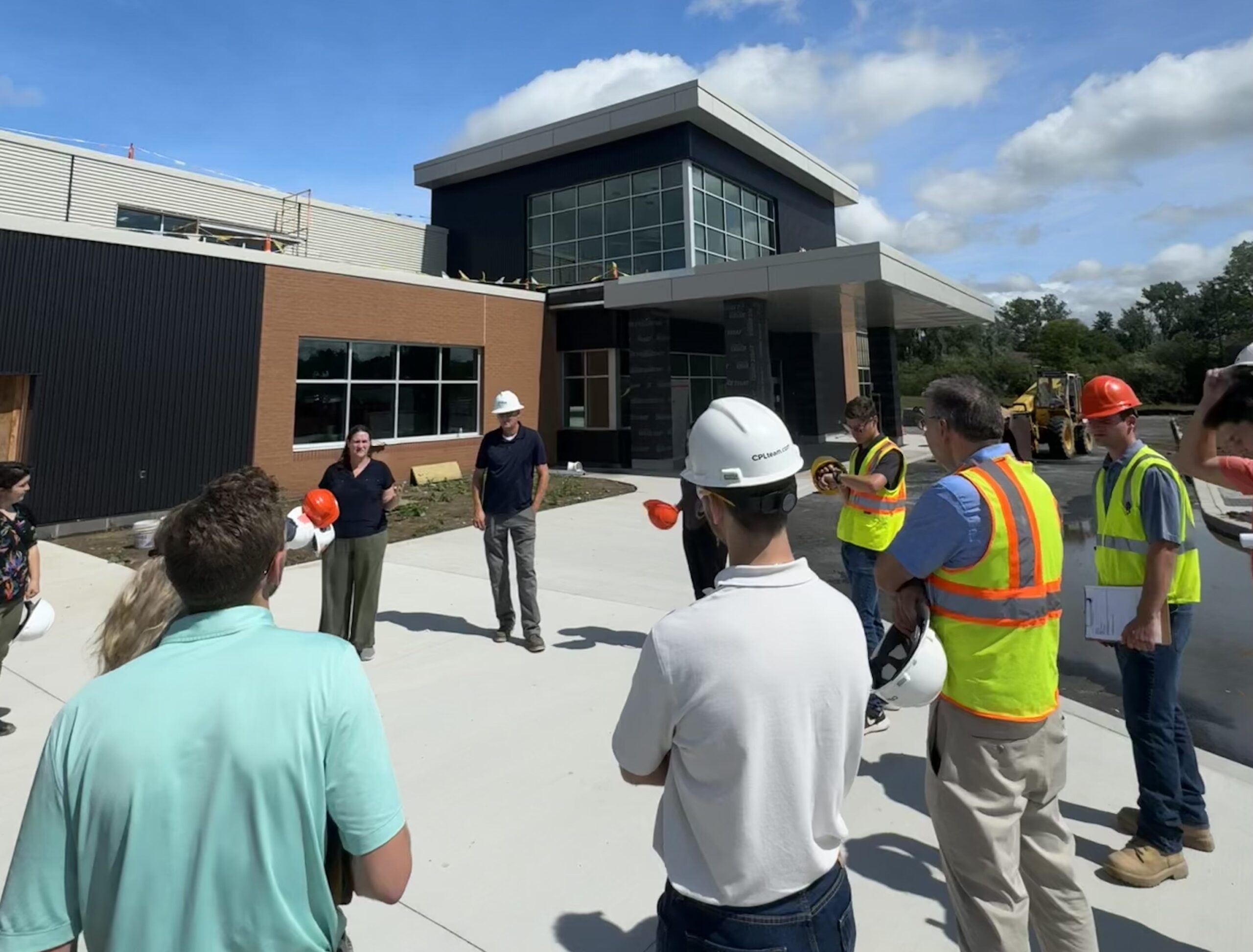 A photo of a group of people visiting a construction site. The building is brick with glass windows, and the sky in the background is blue with white clouds.