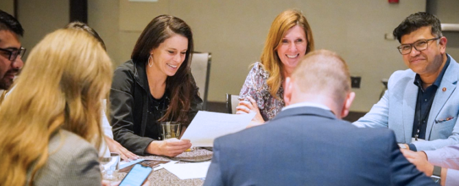 A group of people sit around a table, smiling and talking while they work.