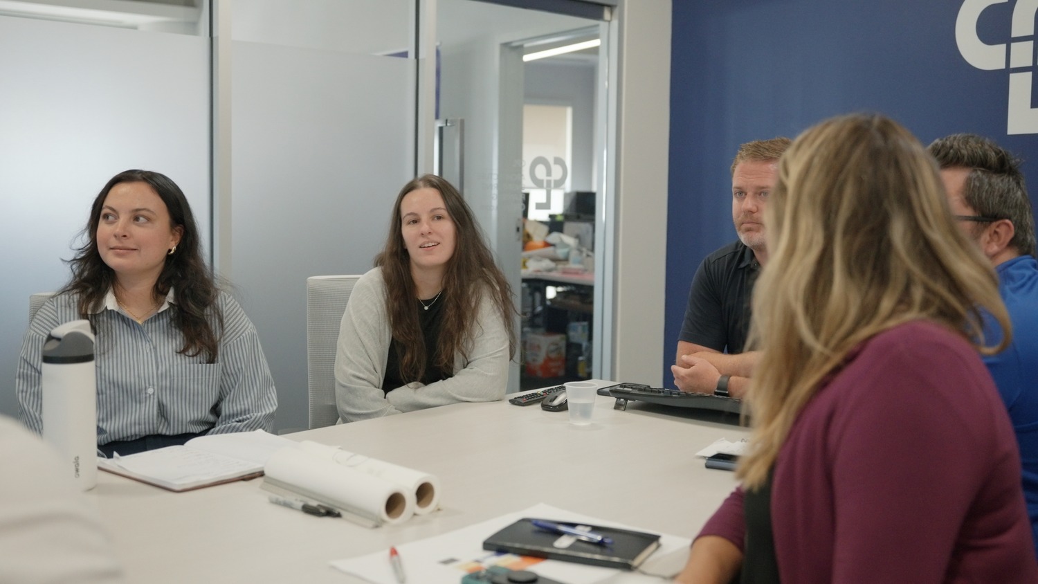 A group of people sit at a white table in a meeting. White drafting paper is at the center of the table, with a blue accent wall in the background.