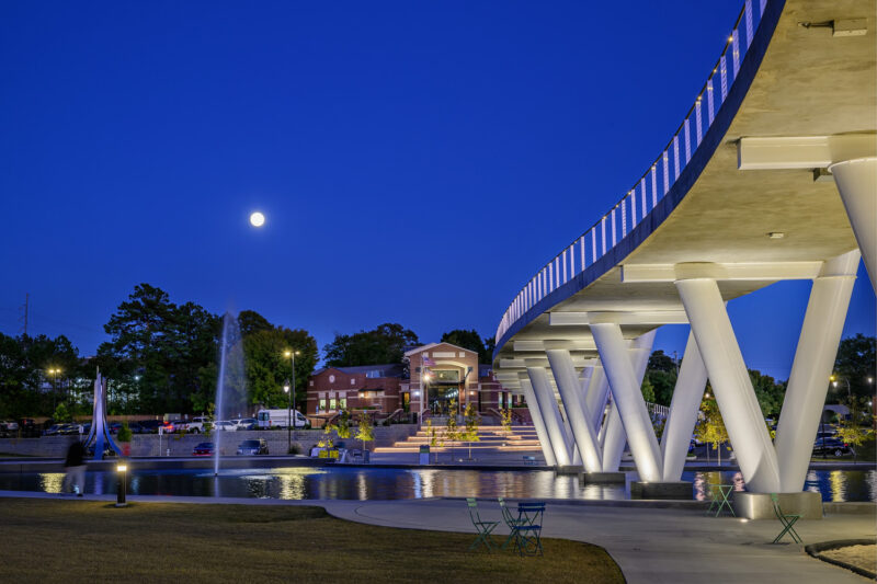 Night view of Suwanee Town Center on Main and DeLay Nature Park. A modern pedestrian bridge arches over the water, supported by white, angular columns. The bridge is illuminated with soft lights, and a full moon is visible in the clear blue sky. In the foreground, a fountain jets water into the pond, with green chairs scattered around the park's paved area. In the background, the red brick building of the town center is visible, surrounded by trees and lit by streetlights.