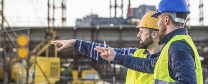 Two men on a construction site wearing neon yellow vests and pointing. One is wearing a yellow hard hat, and the other a blue hard hat. The sky in the background indicates that it is morning.