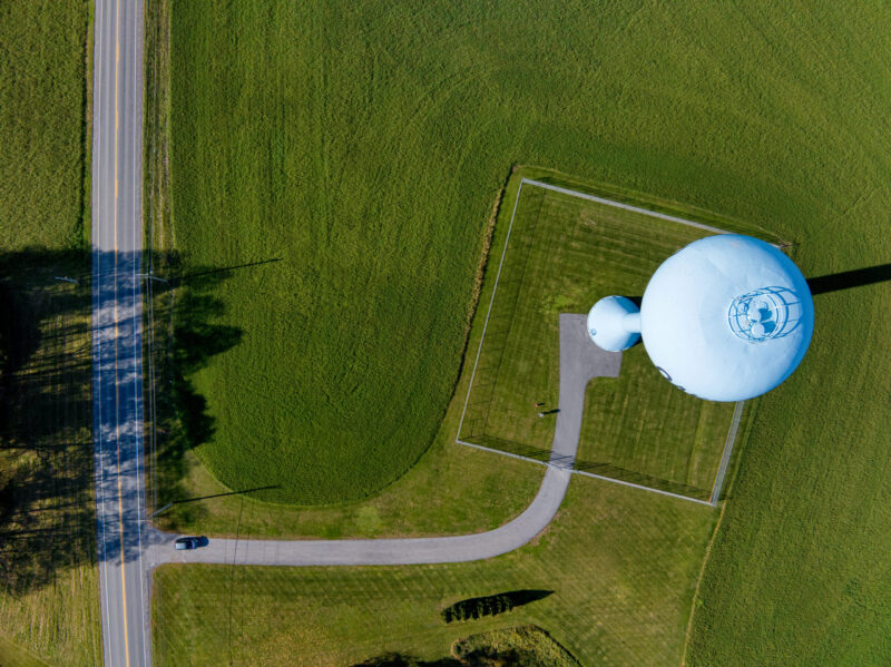 Aerial view of a blue water tower on a vast green field. A road with a black car runs along the left, creating a serene, orderly landscape.