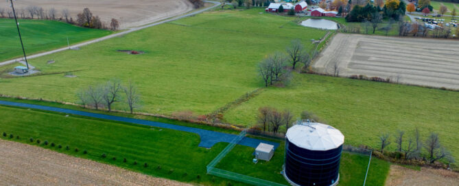 An aerial view of patches of farmland and grass, inluding a water storage tank, and forest in the background.