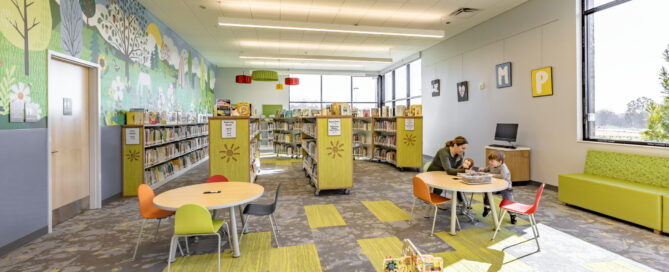A vibrant K-12 library at the Cabarrus Library and Active Living Center. The rug includes colorful green and gray carpeting, with bright lights above and open windows.