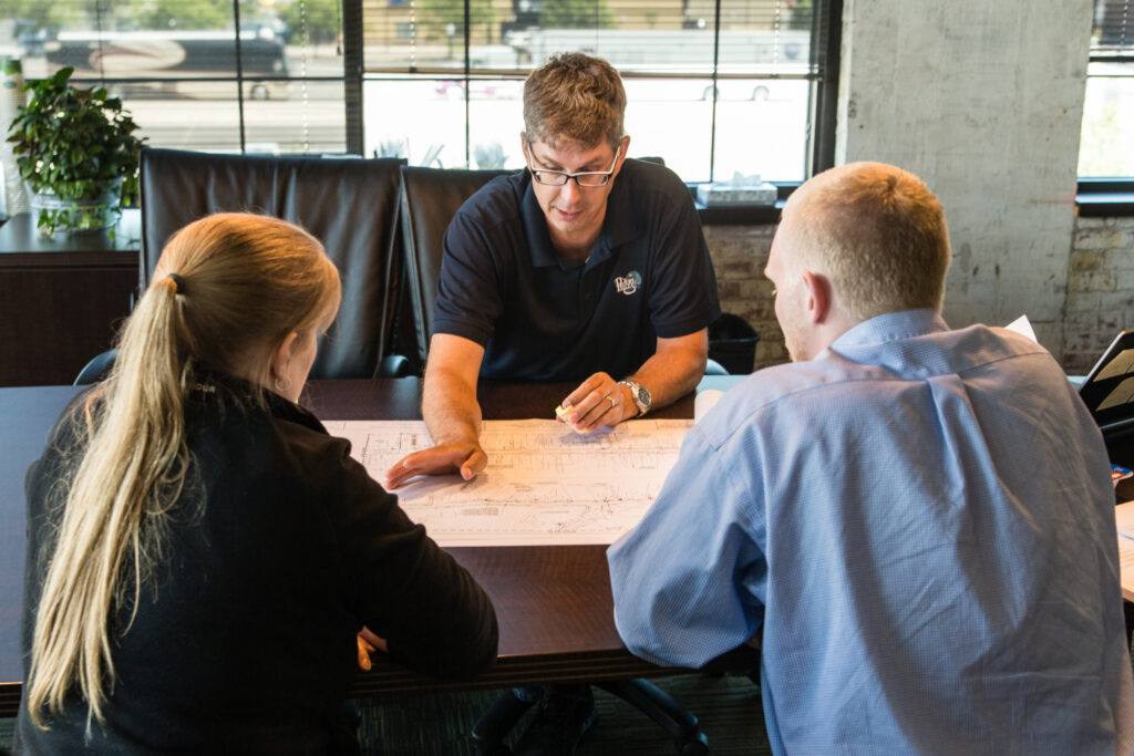 A photo of three people sitting together at a table. One man, CPL National Infrastructure practice leader Eric Wies, in a polo is pointing at a set of plans on a wooden desk, seemingly discussing strategy.