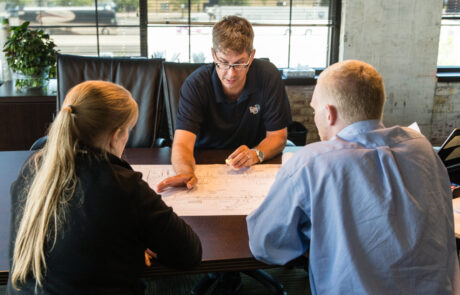 A photo of three people sitting together at a table. One man, CPL National Infrastructure practice leader Eric Wies, in a polo is pointing at a set of plans on a wooden desk, seemingly discussing strategy.