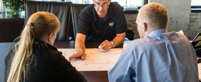 A photo of three people sitting together at a table. One man, CPL National Infrastructure practice leader Eric Wies, in a polo is pointing at a set of plans on a wooden desk, seemingly discussing strategy.