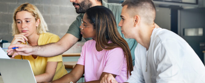 A teacher helping three college students, pointing to something on a document while they observe.