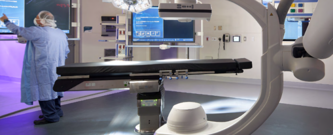 A person wearing scrubs and a face mask stands in a surgical suite next to an operating table, interacting with a touchpad smart screen device.