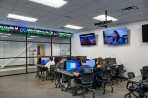 Students in a business hub, typing at rows of desks with computers, and a stock ticker display above glass windows into the room.