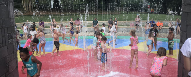Children playing at the center of the splash pad, which has several water features and a soft red surface.