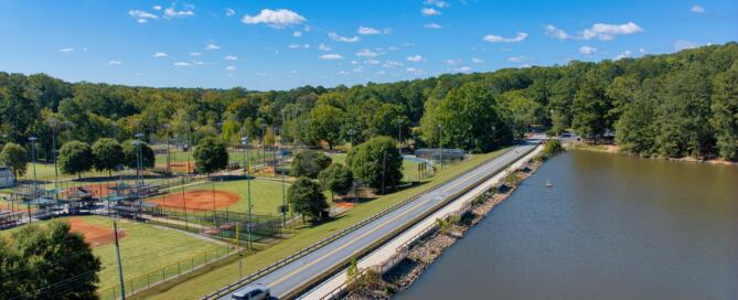 Aerial view of Murphey Candler Park featuring baseball fields, tennis courts, and a roadway running alongside the lake under a clear blue sky.