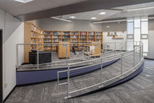 A ramp and guiderails leading to an elevated space in the Odyssey High School library, which has bookshelves lining the walls, chairs, tables, and additional shelves.